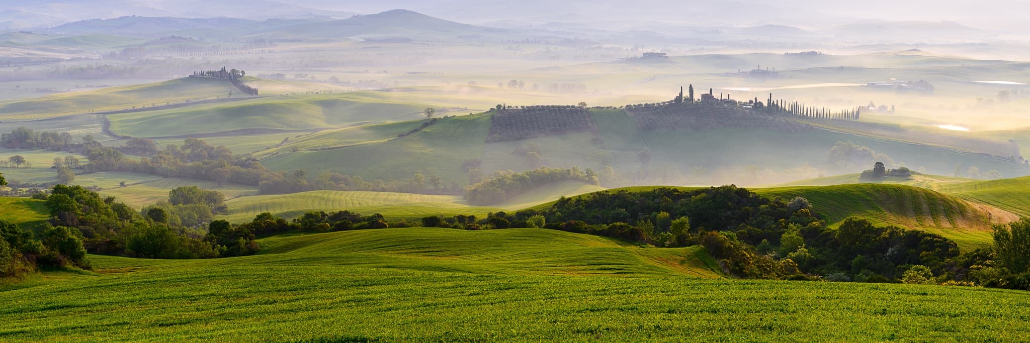 Sunrise over the rolling hills of the Val d'Orcia in Tuscany, Italy — cypress trees on the horizon, morning mist in the valleys between the slopes, International. Photograph by Marty Quinn