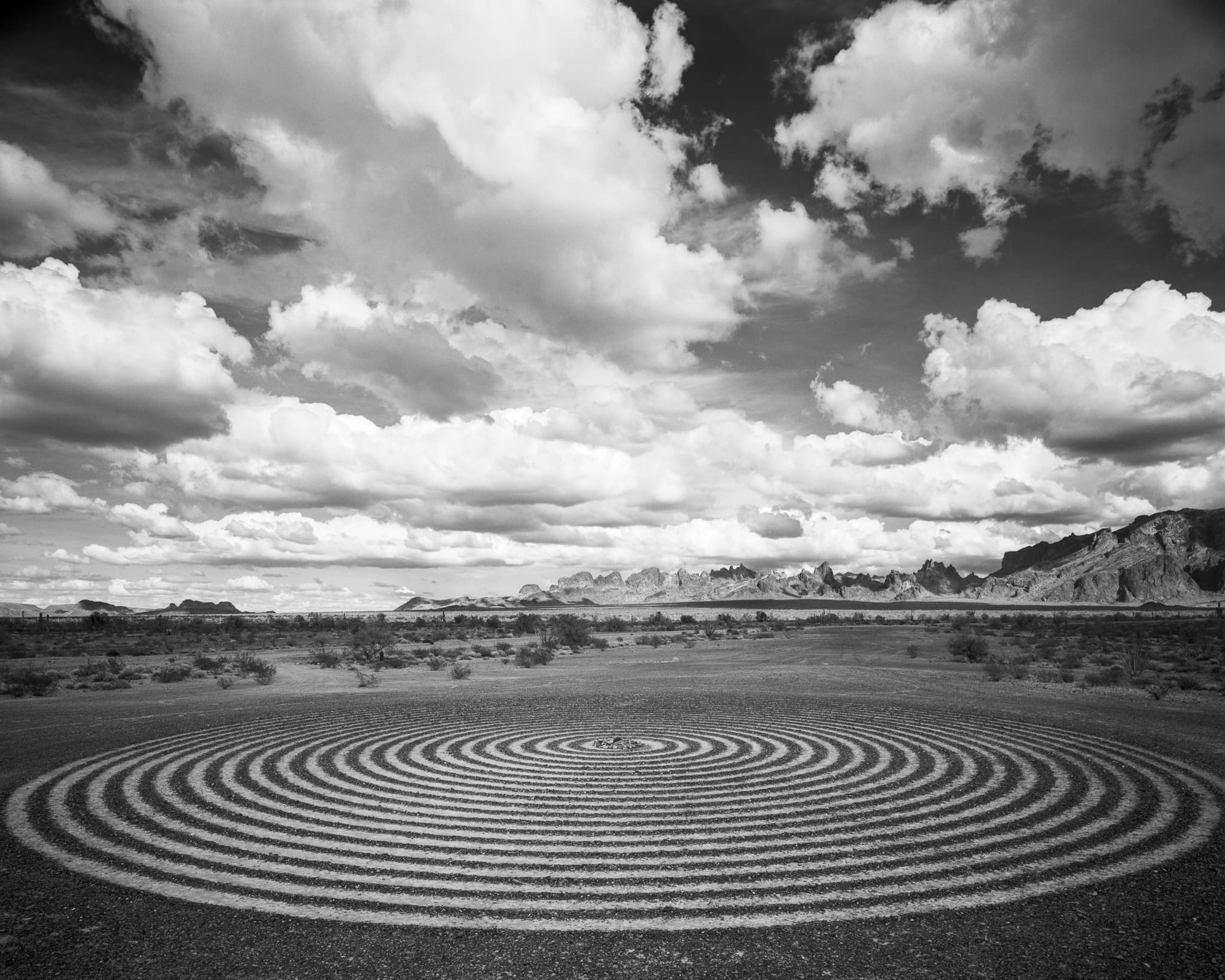 A rock labyrinth laid out across the Mojave desert floor locks eyes with a sky full of cumulus clouds in this large format black and white landscape, Arizona. Photograph by Marty Quinn